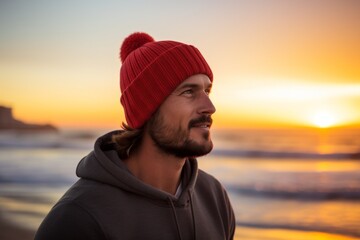 Portrait of a tender man in his 30s donning a warm wool beanie in front of vibrant beach sunset background