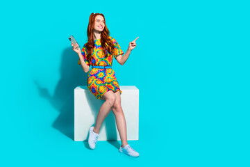 Full size photo of young girl sit cube hold device look point empty space wear colorful outfit isolated on aquamarine color background
