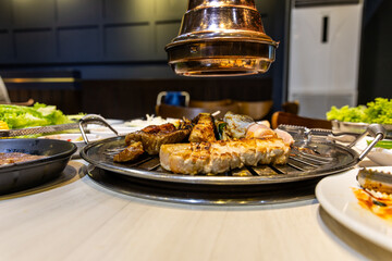 Side view of delicious korean barbeque meal with pieces of beef, pork, chicken placed onto mesh to grill, with condiments and vegetables in restaurant.