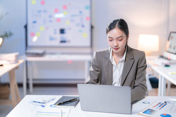 Young professional is concentrating on her laptop, surrounded by charts and documents,...