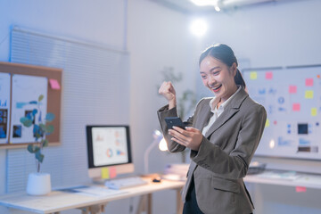 Fototapeta premium Young businesswoman is holding a smartphone and raising her fist, celebrating success after receiving good news on her phone