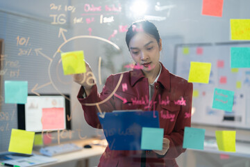 Young businesswoman is sticking adhesive notes on a glass wall while working on a new project. She is holding a clipboard and analyzing data