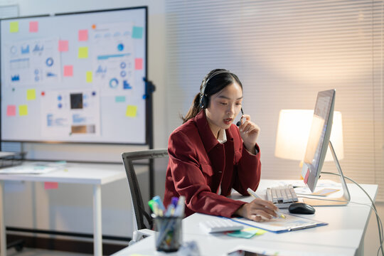 Asian businesswoman working in the office late is wearing a headset and talking on a conference call while analyzing financial charts
