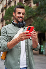 Young caucasian man holding mobile on the street in the city. Happy male wearing headphones and using cell outdoors, looking and checking his social media. Vertical image with copy space.