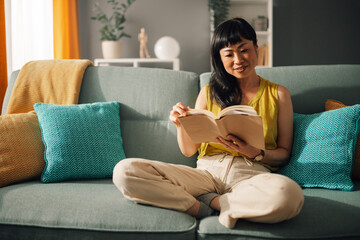 Asian woman reading comfortably on sofa with colorful cushions.