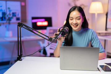Asian woman recording podcast using condenser microphone and laptop computer. She is smiling and having fun in her home studio