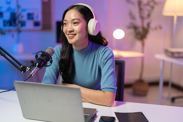Young asian woman is recording a podcast from home, talking into a microphone and using a laptop. She is smiling and looking away from the camera