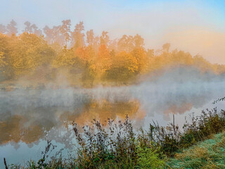 Naklejka premium Sonnenaufgangsstimmung im Herbst. Der Morgennebel liegt über dem Wasserlauf und die Sonne strahlt das bunte Herbstlaub an.