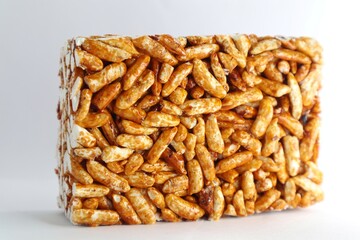 A close-up of a rectangular block of puffed rice treats. The treats are a golden brown color and are coated in a sticky, sweet substance. The block is sitting on a white background.