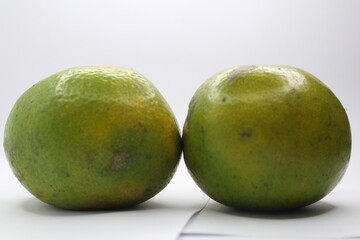 The photo shows two green oranges on a white background. The oranges are round and have a smooth skin. They are slightly different in size.