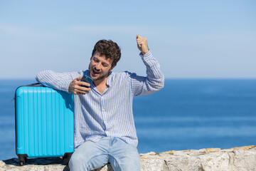 Happy single young adult man booking vacation hotel with his smartphone, blue travel suitcase with...