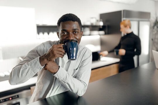 African american man enjoying coffee in modern office kitchen, showcasing relaxed work environment. colleague in background adds to contemporary office scene.