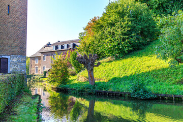 Wijnandsrade Castle moat between church and hill with leafy trees and green grass, reflection on...