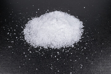 A close-up of a mound of white salt crystals scattered on a dark surface. The textured granules...