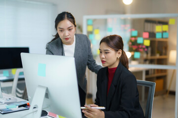 Two asian businesswomen are discussing over a desktop computer, analyzing financial charts and graphs displayed on screen