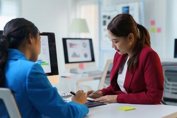 Two young businesswomen wearing suits are reviewing financial data and analyzing charts on a digital tablet in a modern office