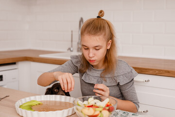 A blonde girl in a grey dress is making an apple pie with a spaniel dog