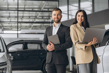 Super sales team in dealership, two consultants or managers in elegant suits with laptop and tablet in arms in car dealership