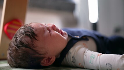 Baby lying on a mat, starting to call for attention with a subtle expression of discomfort. infant...