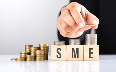 Hand stacking coins with 'SME' written on a wooden block and more coins on the table. Represents financial growth of small and medium-sized enterprises, emphasizing investment and money management.