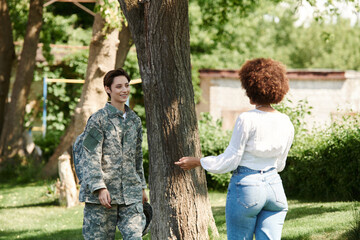 A joyful reunion unfolds as a soldier greets her wife beneath the shade of a tree.