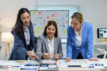 Three asian businesswomen working together, leaning over a desk covered with financial charts and using markers to highlight key data