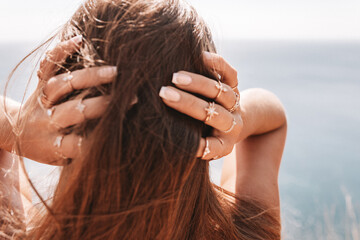 A woman with long brown hair and a beach setting. She is wearing a lot of jewelry, including rings and bracelets.