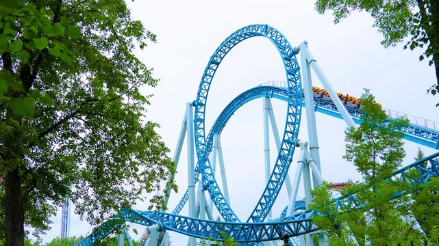 Roller coaster cart with people riding on it goes through an upside down curtain loop, flip, overload. View from among behind trees, bushes, leaves