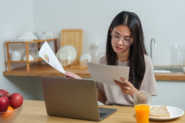 Asian woman sitting at kitchen table reviewing work documents with laptop. Breakfast and orange...