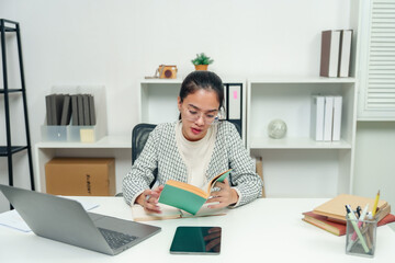 Young Asian woman working in an office setting with a laptop and books. Looking focused and slightly stressed, multitasking with reading and taking notes. Office environment, modern workspace.