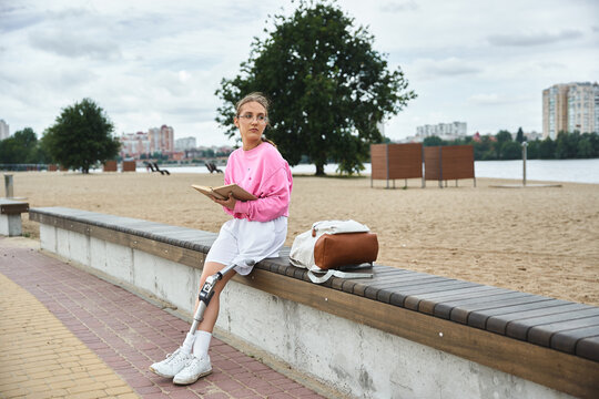 A young woman in sportswear embraces the outdoors, reading by the beach with her prosthetic leg. - Powered by Adobe