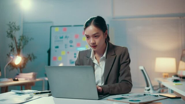 Young asian businesswoman is working late in the office. She is sitting at her desk and typing on her laptop. She is concentrating on her work and is determined to get the job done