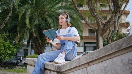 Fototapeta premium Trendy teenager reading book wearing headphones on stone ledge urban park. 