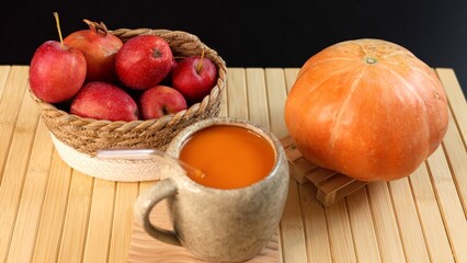 Basket filled with red apples beside ceramic mug with pumpkin beverage, pumpkin placed on bamboo mat, autumn harvest scene showcasing fresh produce, natural textures, and warm seasonal tones.