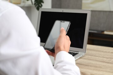 Businessman with smartphone using laptop at wooden table, closeup. Modern technology