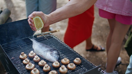 Hand squeezing lemon grilling fish with mushrooms at summer picnic closeup.