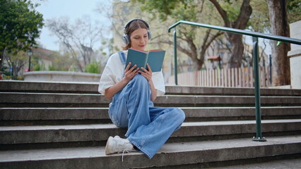 Fototapeta premium Headphones girl enjoy literature sitting on concrete stairs park. Woman reading
