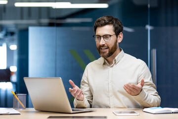 A young successful businessman man is sitting in the office at the work table and is drawing on the video conference online, gesturing with his hands