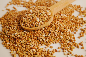 On a white kitchen table, organic buckwheat is piled up, topped with a brown wooden spoon containing some buckwheat.
