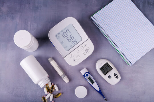 Overhead view of health monitoring tools, including a digital blood pressure monitor, glucose meter, pill bottles, and a notebook on a concrete table