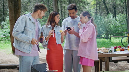 Group friends bonding barbecue at forest picnic. Smiling man showing smartphone