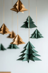 Obraz premium New Year tree decorations made of origami paper in a green and gold color palette hang in front of a white wall adorned with small origami trees.Minimal creative winter holiday concept.