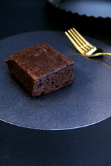 Brownie dessert and fork with baking tray at the back on black background. Dessert table setting of chocolate brownie with dark theme. Sweet, homemade baking, eating, kitchen, and chocolate concepts.