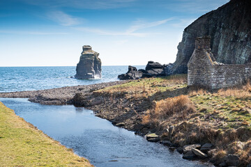 The Burn of Latheronwheel flows into the sea past the ruins of an abandoned croft, In the middle there is a rocky outcrop in the sea reaching up into a blue sky © Glyn