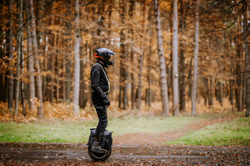 A person wearing a helmet and reflective gear rides an electric unicycle on a forest path surrounded by tall trees and autumn foliage. © Raivo