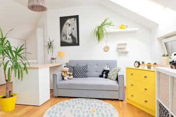 Child room in modern design in the attic with window. Desk with chair and sofa with yellow decor in cozy interior for children. Carpet on wooden floor. Scandinavian style.