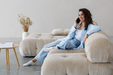 Fototapeta premium Happy beautiful woman is talking on a phone sitting on a sofa at home.