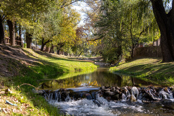 Tranquil stream of water across Alta Gracia town surrounded by grass and trees with horse grazing in autumn