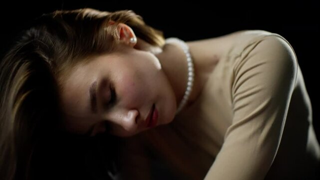 Young woman in a beige leotard and pearl necklace poses elegantly against a black backdrop, displaying fluid movements and captivating expressions