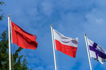 Flags of China, Poland, and Finland waving proudly under a bright blue sky on a sunny day in a peaceful outdoor setting